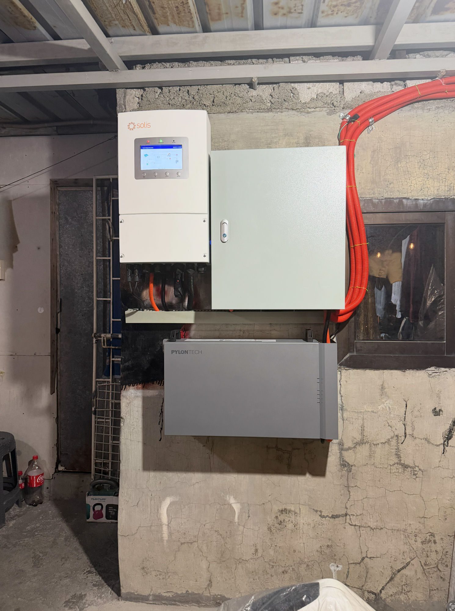 A hybrid inverter, a protection box and a wall battery mounted on a garage wall, orange conduit running up to the roof.